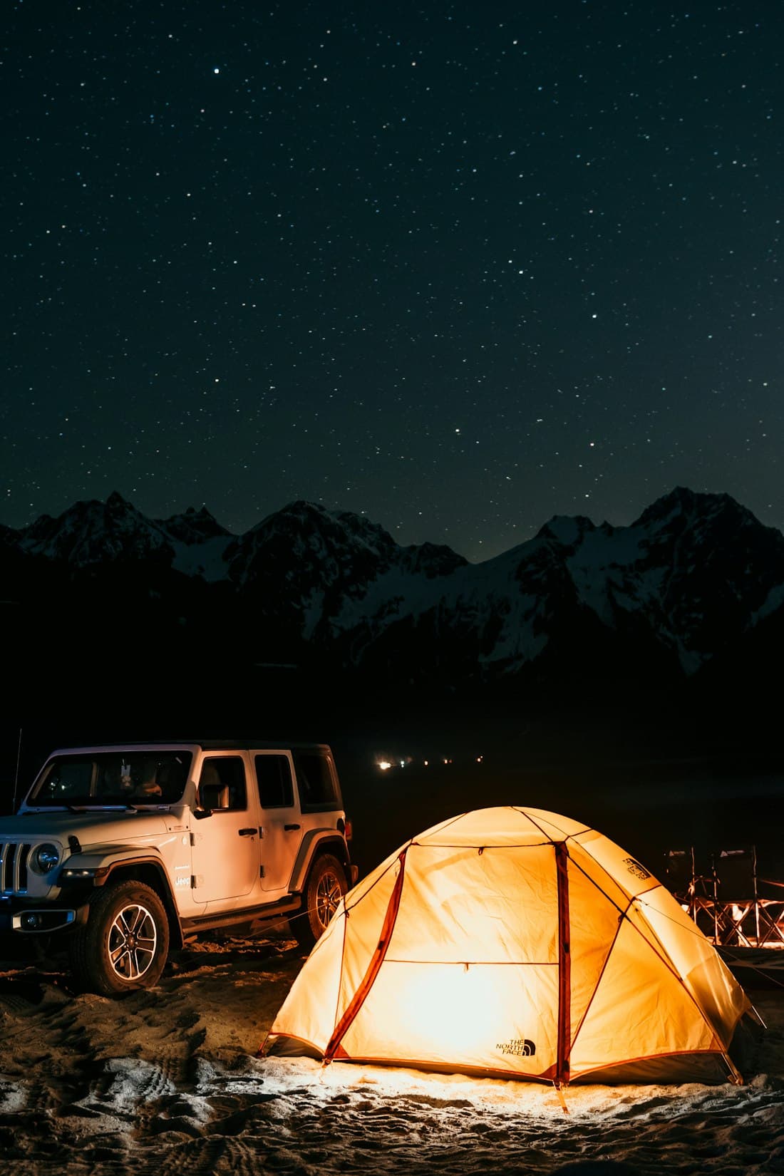 A lit tent and vehicle under a starry night sky with snow-capped peaks
