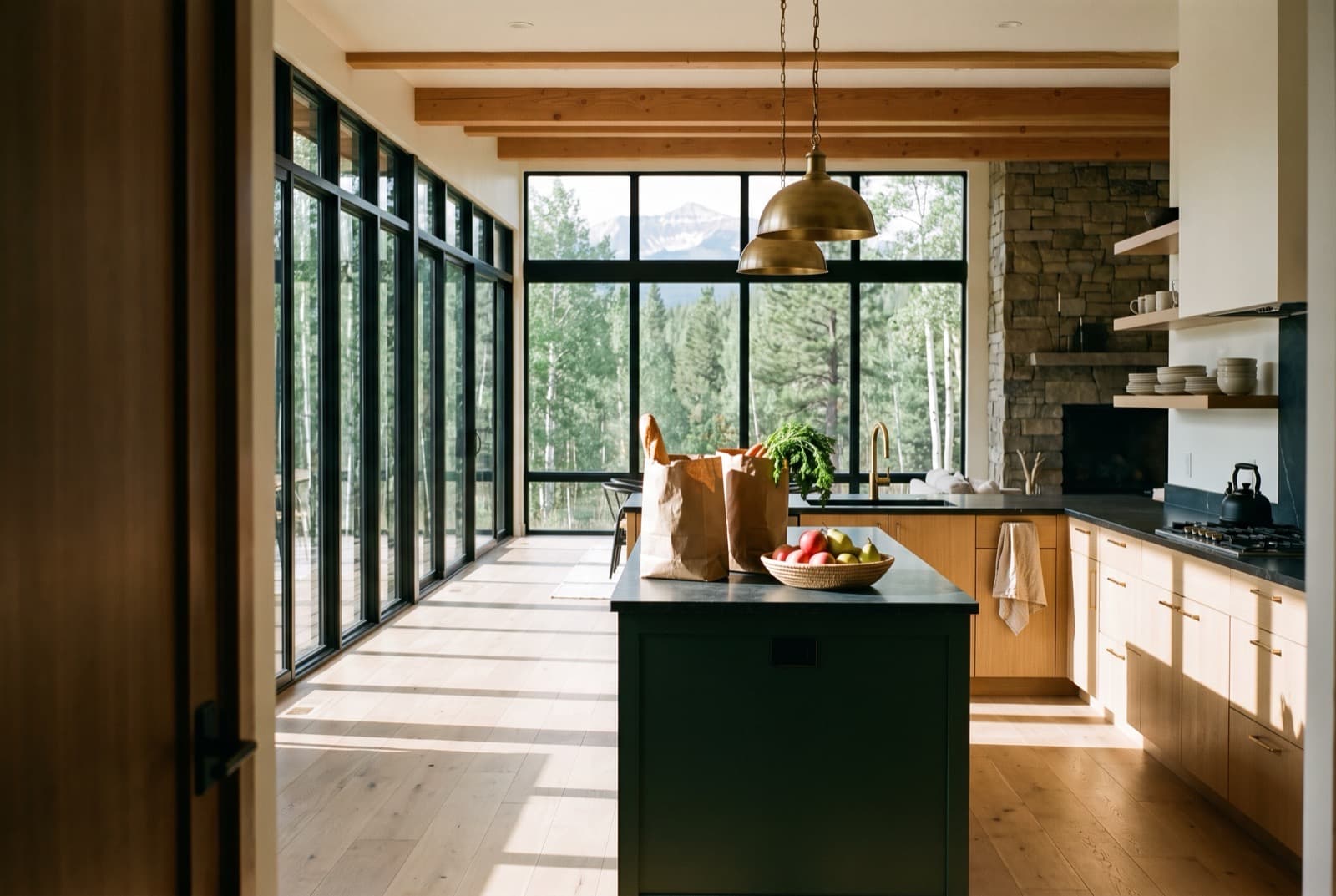 A mountain-modern kitchen and great room with grocery bags and a fruit bowl on the island, mountains through the window wall